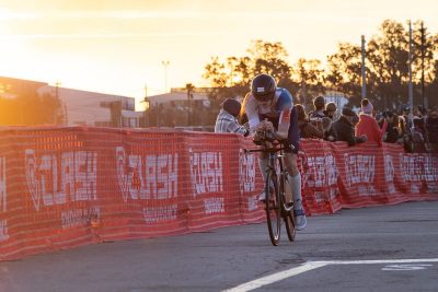 Tommy Doubleday bicycles on a race track, along orange CLASH Daytona signage. A crowd of people stands behind the signage, watching the race. The sun sets in the background.