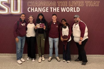 Six people stand smiling before a wall display reading "Changing the Lives That Change the World."