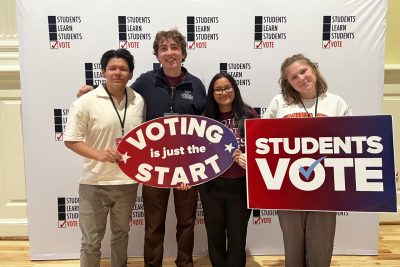 A group of smiling young adults stands holding signs reading "voting is just the start" and "students vote" in front of a Students Learn Students Vote organization-branded backdrop.