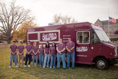 Nine smiling individuals in maroon T-shirts stand in front of a maroon truck labeled "The Chillfield."