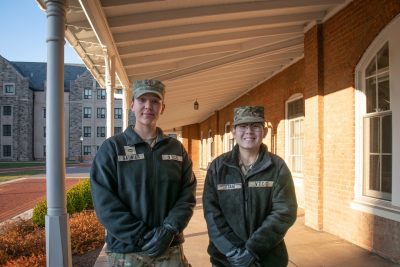 Saunier and Otani stand smiling in uniform under the covered porch of a historic brick building with a Hokie Stone building in the background. 