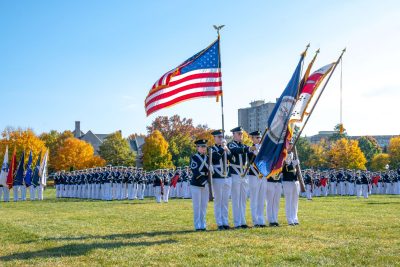 Five uniformed cadets stand in the foreground holding the American flag, the Virginian Flag, and the Corps of Cadets flag, among others. In the background are dozens of rows of more uniformed cadets, all standing at attention. Behind them, you can see trees, the skyline of campus, and a clear blue sky.