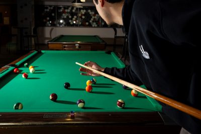 A low-lit image of a pool table, with a person lining up their shot with their back to the camera. The space is low-lit, with the balls casting long shadows on the table.