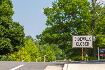A large white sign with bold black letters reading “Sidewalk Closed” is positioned at the edge of a sidewalk next to a road lined with green trees. 