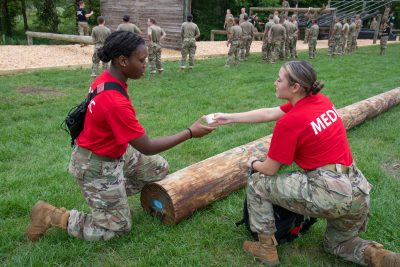 Two people in camo pants and red shirts reading "medic" exchange supplies while several people in the background in full camouflage face an obstacle course.