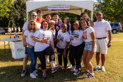 Several adults of mixed ages stand together, smiling on a lawn while dressed in Virginia Tech colors. Some of their shirts read "The Gobble Squad," matching the text a banner in the backbround.